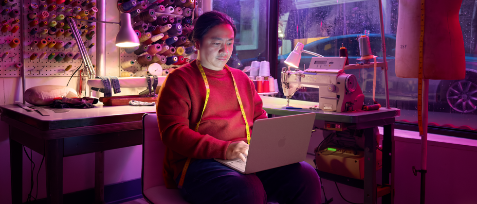 A person uses MacBook Neo, blush color, unplugged on their lap, in a workspace with a sewing machine and related materials, including thread and a dress form